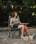 Person sitting in Comfort Pro Rocker Outdoor Rocking Chair with built-in beverage holder, breathable mesh back, at campsite.