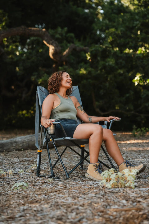 Person sitting in Comfort Pro Rocker Outdoor Rocking Chair with built-in beverage holder, breathable mesh back, at campsite.