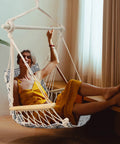 Person relaxing in an upgraded hammock chair with wooden armrests and steel spreader bar, cotton woven fabric, indoor use.