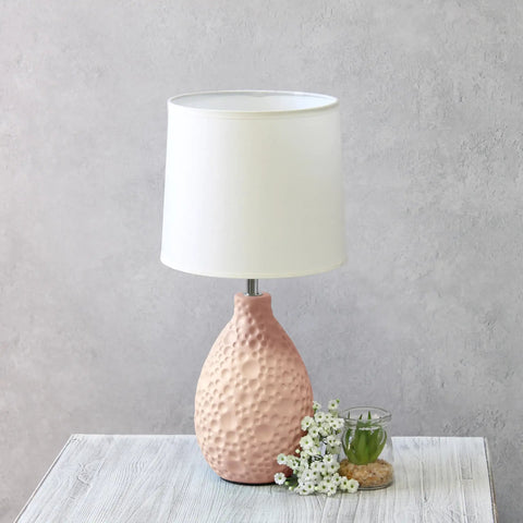 Pink textured ceramic oval table lamp with white fabric shade on a light wood table, decorative plants nearby.