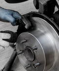Front view of a silver brake rotor with caliper and wheel studs; technician wearing blue gloves