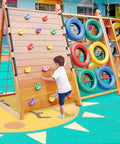 Child climbing wooden playground wall with colorful rock grips next to tire obstacle course on outdoor play area