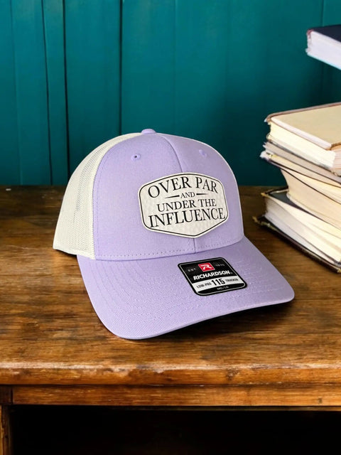 Over Par And Under The Influence Golf Trucker Hat For Women With Dimpled Ball Patch Purple Lilac Birch Color 6 Panel Structured Richardson 115 Cap Funny Golf Accessory displayed on wooden surface