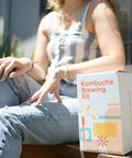 Person sitting with Organic 1 Gallon Kombucha Starter Kit With SCOBY Fermentation Jar Tea And Sugar Bottling Essentials And Clear At Home Brewing Instructions For Beginners To Kickstart Your Brew Like A Pro box.