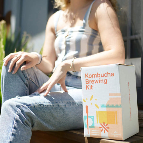 Person sitting with Organic 1 Gallon Kombucha Starter Kit With SCOBY Fermentation Jar Tea And Sugar Bottling Essentials And Clear At Home Brewing Instructions For Beginners To Kickstart Your Brew Like A Pro box.
