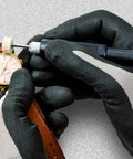 Close-up of hands wearing gloves adjusting a gold watch on a red leather strap with a tool on a gray surface.