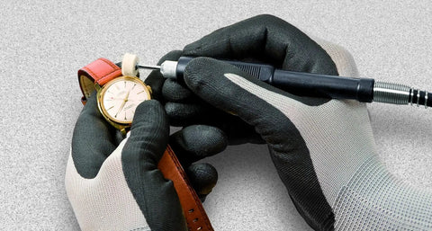 Close-up of hands wearing gloves adjusting a gold watch on a red leather strap with a tool on a gray surface.