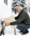 Beige insulated backflow cover installed on outdoor irrigation valve; man kneels in snow beside house.