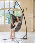 Woman sitting in Hammock Chair With Stand For Indoor And Outdoor Use, Chill Egg Swing Chair With Patented Adjustable Pillow, Two Cushions, Side Pocket, Bag Included, 350 Lb Capacity, Dark Gray, placed indoors by large windows.