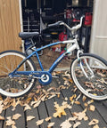 Blue comfort bike with white tires beside a shed on a wooden platform surrounded by fallen leaves