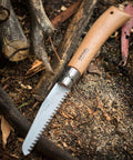 Badass Lightweight Folding Saw For Weekend Campouts And Garden Grind Cuts Branches Fast With Razor Sharp Carbon Steel Blade Secure Virobloc Lock And Natural Beechwood Handle on wooden ground with sticks.