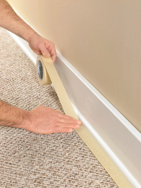 Tan Masking Tape 0.75 Inch By 60 Yard General Purpose Tape For Paint Masking Bundling And Mounting Ideal For Home Workshop And DIY Projects Pack Of 12 being applied along baseboard on carpeted floor.