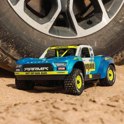 Grom 4x4 Desert RC Truck Ready To Run With Battery And Charger Included 2S LiPo Smart Charger All Terrain Enclosed Metal Gear Drivetrain Beefy V2 Differentials Clipless Body Mojave Style Blue White on sand next to tire