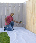 A person using the No Fuss Heavy Duty Airless Paint And Stain Sprayer on a wooden fence, kneeling on a protective tarp.