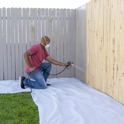 A person using the No Fuss Heavy Duty Airless Paint And Stain Sprayer on a wooden fence, kneeling on a protective tarp.