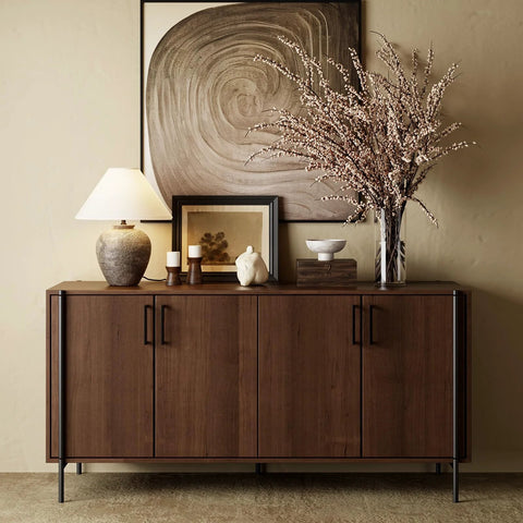 Wooden sideboard with dark square cabinet handles, decorative lamp, artwork, and floral arrangement on top.