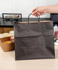 100 Pack Stand Open Paper Shopping Bags With Handles For Grab And Go Takeout And Retail Durable Kraft Paper Carrier Bags 14.17 X 8.26 X 13.19 Inches Easy Carry Totes For Gifts Groceries And Everyday Shop Runs shown in use on a countertop