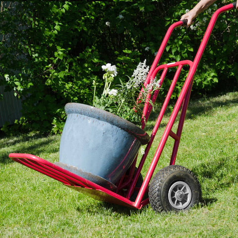 Red hand truck with 10x3.00-4 flat free tire and wheel assembly carrying large planter, showing side view on grass