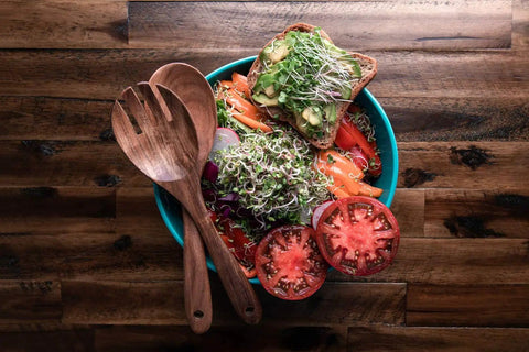 Fresh salad with microgreens, tomatoes, toast, and wooden utensils on a wooden table, featuring Colorful Microgreen Seeds Mix for Home Growers.