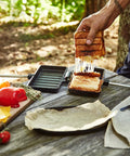 Hand lifting grilled sandwich from XL Cast Iron Pie Iron Sandwich Maker For Campfire Cooking on outdoor table.