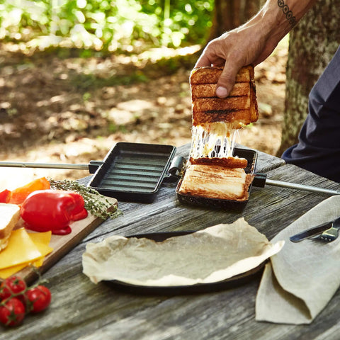 Hand lifting grilled sandwich from XL Cast Iron Pie Iron Sandwich Maker For Campfire Cooking on outdoor table.
