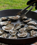 Oysters cooking in a cast iron pan outdoors, ready for the Premium Stainless Steel Oyster Opener with Knife on Wooden Base