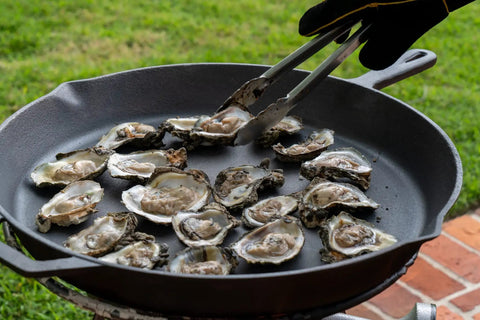 Oysters cooking in a cast iron pan outdoors, ready for the Premium Stainless Steel Oyster Opener with Knife on Wooden Base