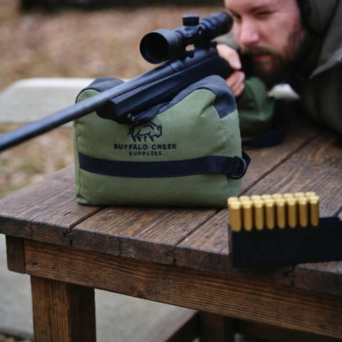 Pre Filled All Weather Rifle Sandbag Rest For Bench Shooting And Competition Front And Rear Set Ready To Use Heavy Duty 900D Oxford Fabric Tear Resistant Olive Drab on wooden table with scope mounted rifle