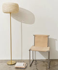 Rowan Brass Floor Lamp With Unique Thread Shade next to a modern beige chair on a concrete floor, mid-century boho decor.