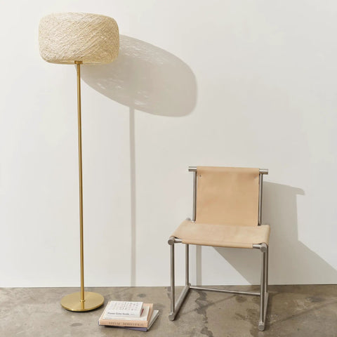 Rowan Brass Floor Lamp With Unique Thread Shade next to a modern beige chair on a concrete floor, mid-century boho decor.