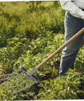 Heavy Duty Steel Garden Fork For Soil Loosening And Deep Soil Turning used in a garden by person wearing gloves and jeans