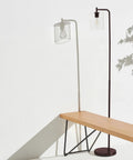 Tall Edison Bulb Floor Lamp With Glass Shade In Bronze Finish next to wooden bench, casting shadow on white wall, indoor setting