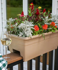 Super Sleek 24 Inch Deck Rail Planter Box With Built In Drainage Plug, UV Resistant Plastic Outdoor Planters And Flower Box For Garden Patio Balcony Fence, Sandstone Finish, Straight Up Sturdy For Any Backyard Space filled with colorful flowers and displayed with a glass of wine.