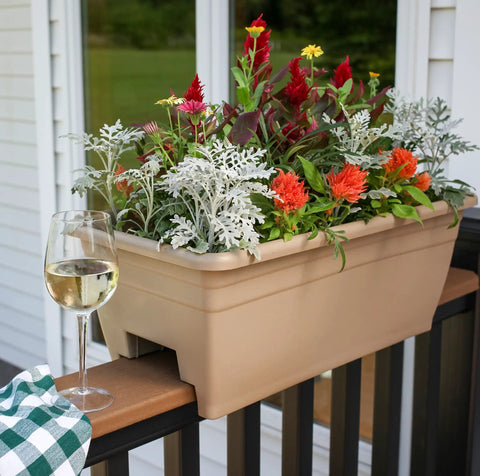 Super Sleek 24 Inch Deck Rail Planter Box With Built In Drainage Plug, UV Resistant Plastic Outdoor Planters And Flower Box For Garden Patio Balcony Fence, Sandstone Finish, Straight Up Sturdy For Any Backyard Space filled with colorful flowers and displayed with a glass of wine.