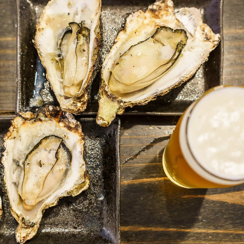 Three grilled oysters on the half shell served on a black plate next to a glass of beer.
