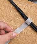 Hand holding a white tearable cable management strap securing black cords on a wooden surface. Heavy Duty Reusable Cable Management Straps White Tearable Cord Organizer Tie For Computer Speaker Guitar Wires Snag Up Messy Cords And Keep Your Desk Looking Super Clean Every Day.