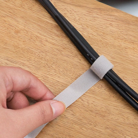 Hand holding a white tearable cable management strap securing black cords on a wooden surface. Heavy Duty Reusable Cable Management Straps White Tearable Cord Organizer Tie For Computer Speaker Guitar Wires Snag Up Messy Cords And Keep Your Desk Looking Super Clean Every Day.