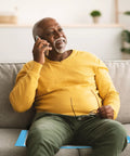 Senior man in yellow shirt sitting on a gray couch talking on phone, smiling pleasantly, while holding glasses.