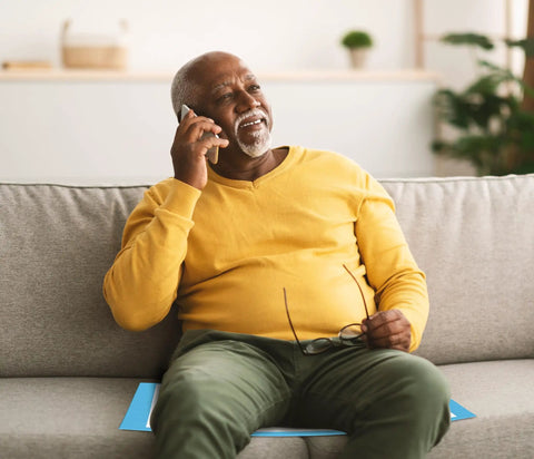 Senior man in yellow shirt sitting on a gray couch talking on phone, smiling pleasantly, while holding glasses.