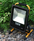 Spot Lighting 50W Led Rechargeable Construction Floodlight, 5000Lm Sup, with sturdy stand on rocky ground surrounded by green foliage.