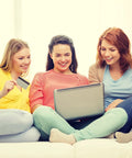 Three women sitting on a sofa smiling while looking at a laptop screen, one holds a credit card, light and casual indoor setting.