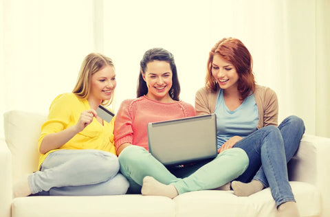Three women sitting on a sofa smiling while looking at a laptop screen, one holds a credit card, light and casual indoor setting.