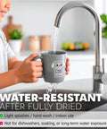 Gray ceramic mug with a cartoon cat sticker being rinsed under a kitchen faucet
