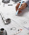 Person drawing mechanical diagrams with a blue pencil on technical papers surrounded by mechanical parts and tools.