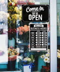 Open Closed Sign Set With Business Hours Flip Sign For Window And Door Includes Vinyl Sticker Pack Eye Catching Open For Biz And Closed Messages Perfect For Store Restaurant Or Bar Black displayed on a glass door with flowers.