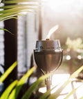 65 Inch Outdoor Torch With Amped Up Extra Large Flame For Patio Garden Lawn And Backyard Weather Resistant Refillable And Decorative Torch In Silver Finish Twist And Pour Refilling For Mess Free Fueling, illuminating a patio in sunlight.