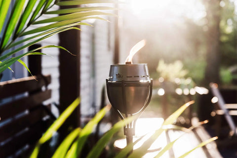 65 Inch Outdoor Torch With Amped Up Extra Large Flame For Patio Garden Lawn And Backyard Weather Resistant Refillable And Decorative Torch In Silver Finish Twist And Pour Refilling For Mess Free Fueling, illuminating a patio in sunlight.