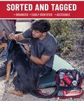 Surviveware 238-piece survival first aid kit bag (red/black) opened beside a man and dog on a campsite.