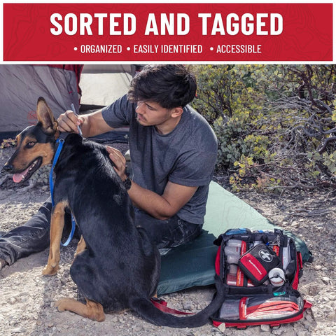 Surviveware 238-piece survival first aid kit bag (red/black) opened beside a man and dog on a campsite.
