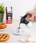Person pouring coffee from Induction Moka Pot That Just Works On Any Hob 4 Cup Espresso Maker 5.7 Oz Black Aluminum Upper With Stainless Steel Boiler Easy Brews On Any Stove Classic Italian Coffee On Demand into a cup next to pastries.